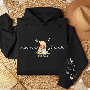 May include: Black hooded sweatshirt with a Winnie the Pooh illustration and the text "mama bear est. 2024". The sleeve has the text "Alex Sarah Lucas" with bee illustrations.