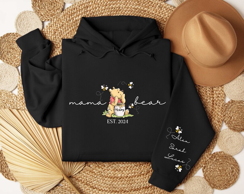 May include: Black hooded sweatshirt with a Winnie the Pooh illustration and the text "mama bear est. 2024". The sleeve has the text "Alex Sarah Lucas" with bee illustrations.