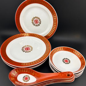 May include: A set of red and gold Chinese porcelain dinnerware with a traditional design. The set includes a large plate, a smaller plate, and a set of soup spoons.