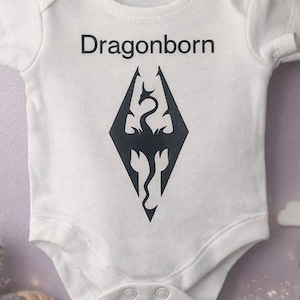 Op de afbeelding: Witte babyromper met het woord "Dragonborn" in zwart gedrukt boven een zwarte ruitvormige afbeelding. De afbeelding toont een gestileerde draak. De romper hangt aan een waslijn met houten knijpers.