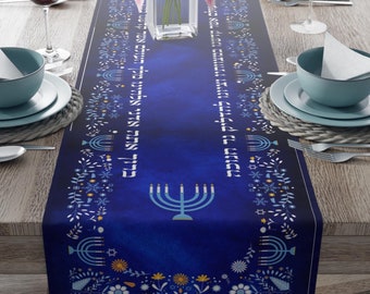 Puede incluir: Camino de mesa azul con diseño de menorá. El camino de mesa presenta un fondo azul oscuro con texto hebreo blanco e ilustraciones de menorá. Patrones florales y de estrellas bordean los bordes. El camino de mesa está sobre una mesa de madera.