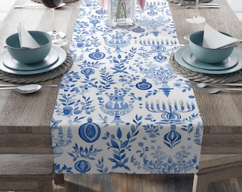May include: A white table runner with a blue floral and ornamental design. The runner features a repeating pattern of stylized leaves, vases, and ornaments. The table is set with blue bowls, plates, and wine glasses, creating a cohesive dining aesthetic.