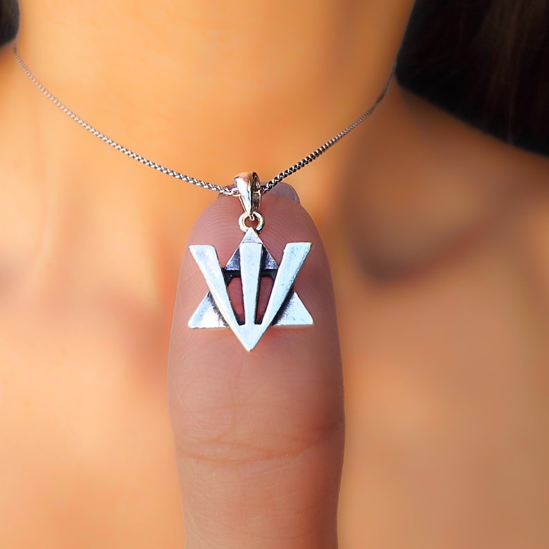 A silver pendant with a triangular symbol is being held by a person's thumb.