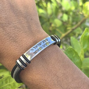 Könnte beinhalten: Ein schwarz-silbernes Armband mit hebräischen Text. Das Armband hat eine silberne rechteckige Platte mit eingravierten hebräischen Buchstaben. Das Band ist schwarz mit silbernen Akzenten und wird am Handgelenk getragen.