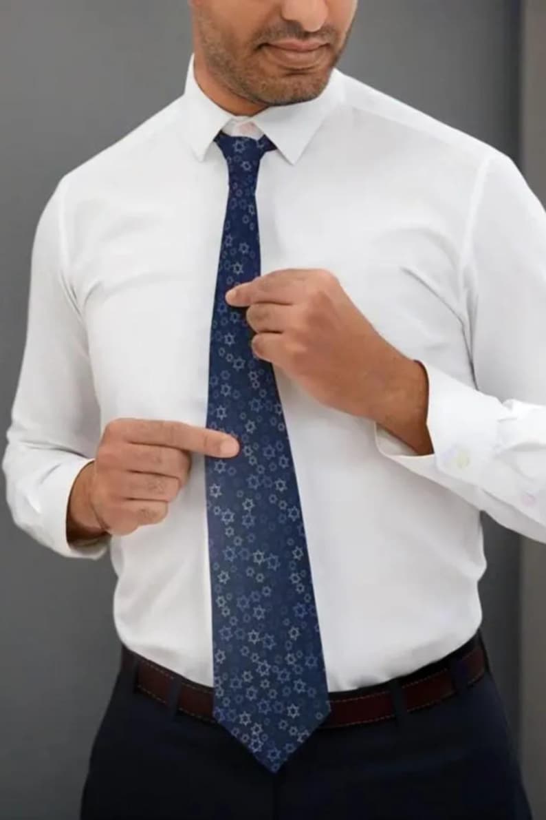 A man in a white shirt and blue tie is adjusting his tie, pointing to a specific part of it.
