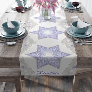 May include: A white table runner featuring a repeating pattern of purple six-pointed stars with intricate floral details and beige geometric accents. The word Weinstein is printed in purple script at the bottom edge of the fabric runner.