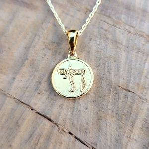 A gold-colored pendant necklace with a circular medallion featuring an image of a letter on it, resting on a wooden surface.
