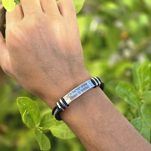 May include: A black leather bracelet with a silver metal plate. The plate has Hebrew text engraved on it. The bracelet has silver accents and is worn on a wrist. The background is blurred green foliage.