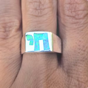 May include: A silver ring with a rectangular face. The face features a Hebrew letter design filled with a vibrant, opalescent blue-green material. The ring is shown on a person's finger.