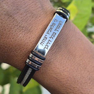 May include: A silver and black bracelet with Hebrew text. The bracelet features a rectangular silver plate with engraved text, attached to a black band with a mesh design. The bracelet is worn on a wrist.