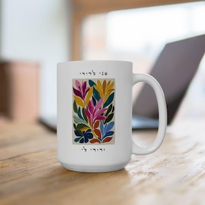 May include: White ceramic mug with a vibrant floral design in pink, yellow, blue, and green. The mug has a large handle and text in Hebrew. The design is a rectangular shape on the front of the mug.
