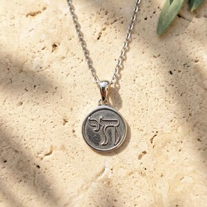 May include: A silver-toned necklace featuring a circular pendant with a textured background and the Hebrew letter Chai embossed in the center. The pendant hangs from a delicate silver-toned chain, creating a simple and classic jewelry piece.