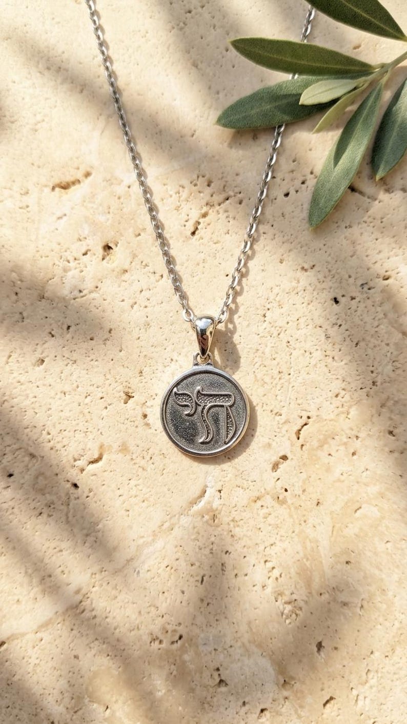 May include: A silver-toned necklace featuring a circular pendant with a textured background and the Hebrew letter Chai embossed in the center. The pendant hangs from a delicate silver-toned chain, creating a simple and classic jewelry piece.