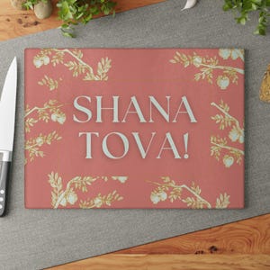 May include: A rectangular cutting board with a coral background and the text "SHANA TOVA!" in light blue. The board is decorated with gold-colored branches and fruit. A knife is visible on the left side.