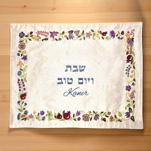 Może przedstawiać: Kremowy prostokątny podkładka z haftowaną kwiatową i granatową obwódką. Tekst "Shabbat Shalom Kaner" jest wyhaftowany na niebiesko. Podkładka jest umieszczona na drewnianej powierzchni.