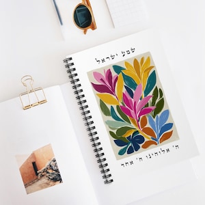 May include: A spiral-bound notebook with a colorful floral illustration in shades of pink, yellow, blue, and green. The notebook is open, revealing a photo and a gold binder clip. Sunglasses and a pen are also visible.
