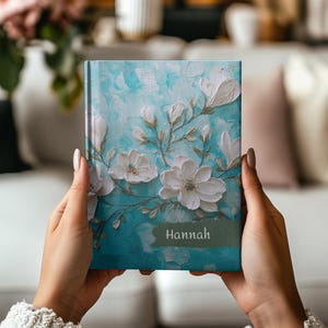 May include: A teal journal with white floral embellishments and the name "Hannah" printed on the cover. The journal features a textured, artistic design with raised white flowers and green stems.
