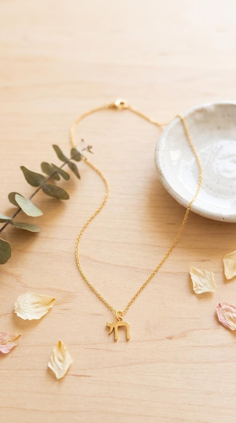 May include: A gold-colored necklace featuring a delicate chain and a pendant shaped like the Hebrew letter Chai. The pendant is suspended from a thin, polished metal chain that includes a small circular clasp mechanism at the back of the necklace.