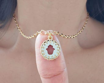 Può includere: Una collana color oro con un ciondolo raffigurante un simbolo della mano di Hamsa. Il ciondolo ha uno sfondo bianco con un disegno a mano rosso e un bordo dorato decorativo. Anche la catena è color oro.