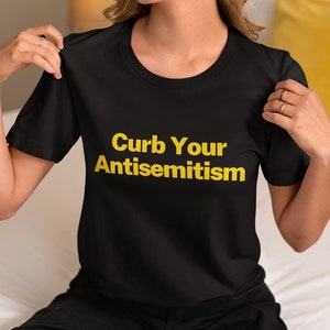 Könnte beinhalten: Schwarzes T-Shirt mit dem Text "Curb Your Antisemitism" in gelben Buchstaben.