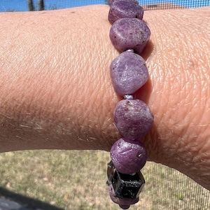 May include: A purple gemstone bracelet with a black gemstone charm. The bracelet is made of round, faceted stones and is strung on a thin, white cord.