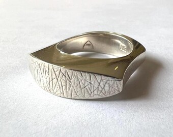 May include: A silver ring with a textured, rectangular design. The ring has a smooth, polished interior and a textured exterior.