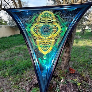 Puede incluir: Un tapiz triangular con un diseño psicodélico que presenta un mandala verde, azul y dorado con patrones geométricos. El tapiz está colgado de una rama de árbol.
