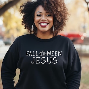 May include: A black sweatshirt with the text "FALL-WEEN JESUS" in white. The word "WEEN" features a pumpkin graphic. The person wearing the sweatshirt is smiling, with curly brown hair and silver hoop earrings. The background is blurred with fall colors.