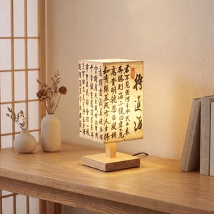 Peut inclure: Lampe carrée avec un socle en bois et un abat-jour orné de calligraphie. La lampe est allumée, diffusant une lumière chaleureuse. Elle est posée sur une table en bois, avec des vases décoratifs et des livres en arrière-plan.