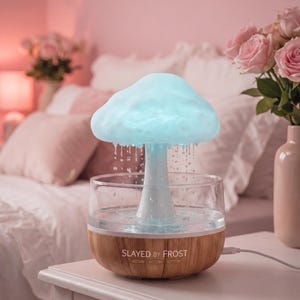 Peut inclure: Diffuseur d'huiles essentielles en forme de champignon avec un sommet bleu clair en forme de nuage et une tige blanche. Le diffuseur est posé dans un bol en verre transparent avec une base en bois. Le texte "SLAYED BY FROST" est imprimé sur la base.