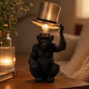 Peut inclure: Lampe de table noire en forme de gorille avec un abat-jour en forme de chapeau haut de forme doré. L'ampoule est placée à la place de la tête du gorille. La lampe est posée sur une surface en bois.