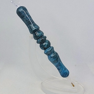 Peut inclure: Stylo en résine bleu et turquoise avec une pointe claire et angulaire et un manche décoratif et arrondi. Le stylo est présenté sur un support en acrylique transparent, mettant en valeur son design marbré. Le design du stylo rappelle les vagues de l'océan.