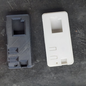 Könnte beinhalten: Zwei 3D-gedruckte Handyhüllen, eine schwarze und eine weiße. Beide Hüllen haben einen rechteckigen Ausschnitt für eine Kameraobjektiv und drei rechteckige Ausschnitte für Tasten.