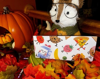 May include: A white card with a colorful pattern of cartoon food characters. The characters include a tomato, a cookie, a strawberry, a pizza, a banana, a carton of milk, a donut, and a pea. The card is sitting on a pile of fall leaves.