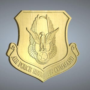 Puede incluir: Una placa de madera de color dorado con el emblema del Comando de la Reserva de la Fuerza Aérea. El emblema presenta un águila estilizada con las alas extendidas, una estrella y un rollo con las palabras "Air Force Reserve Command".
