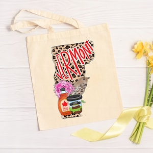 May include: A beige tote bag with a design featuring the state of Vermont in a leopard print with the word "Vermont" in red text. The design also includes a maple syrup bottle, a coffee cup, a bird, and pink flowers.