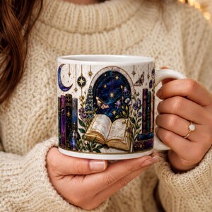 A person is holding a mug with a night sky and stars design, featuring a book and other celestial elements.