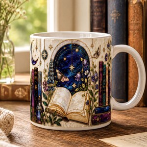 A white mug with a detailed illustration of a book, flowers, and a starry night sky on a wooden surface, with a vase of white flowers in the background.