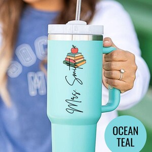 May include: A teal insulated tumbler with a white lid and handle. The tumbler features a stack of books with an apple on top and the text "Mrs. Smith" in a script font. A teal circle with the words "Ocean Teal" is in the bottom right.