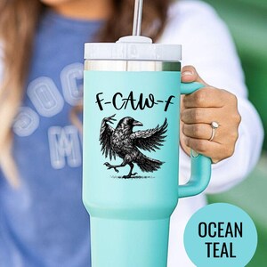 A stainless steel insulated tumbler featuring a detailed black raven illustration with wings spread, paired with the phrase "F-Caw-F" in playful script; the design blends gothic and humorous elements, creating a unique travel cup for bird enthusiasts, crow lovers, or fans of dark sarcastic quotes; shown on tumblers in various matte finishes, each with a handle and straw for practical everyday use.