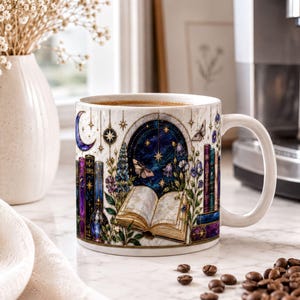 A white mug with a colorful illustration of a book, flowers, and a crescent moon on a white countertop, with coffee beans scattered around it.