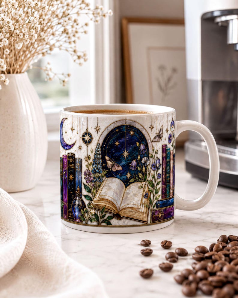 A white mug with a colorful illustration of a book, flowers, and a crescent moon on a white countertop, with coffee beans scattered around it.