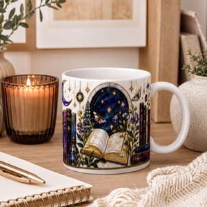 A white mug with a colorful illustration of a book, flowers, and a starry night sky, placed on a wooden surface next to a notebook, a candle, and some books.