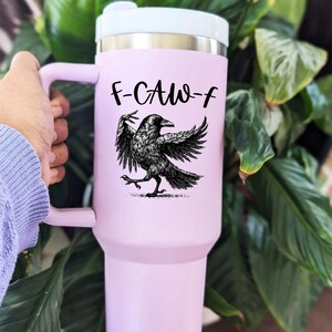 A stainless steel insulated tumbler featuring a detailed black raven illustration with wings spread, paired with the phrase "F-Caw-F" in playful script; the design blends gothic and humorous elements, creating a unique travel cup for bird enthusiasts, crow lovers, or fans of dark sarcastic quotes; shown on tumblers in various matte finishes, each with a handle and straw for practical everyday use.