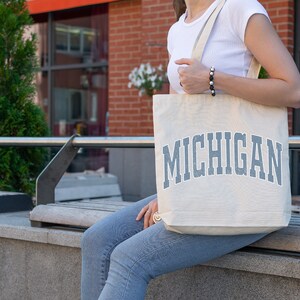 May include: A person carrying a white canvas tote bag with the word "MICHIGAN" printed in blue on the front.