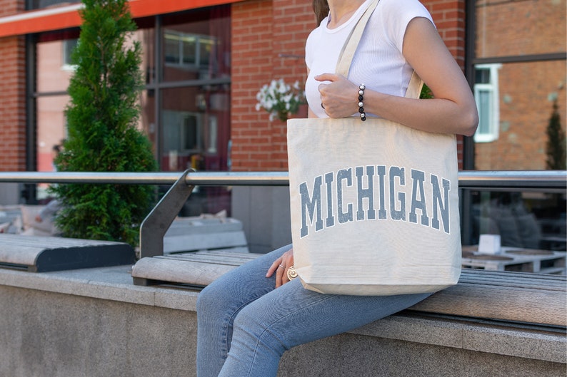 May include: A person carrying a white canvas tote bag with the word "MICHIGAN" printed in blue on the front.