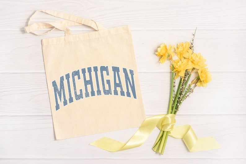 May include: A beige tote bag with the word "MICHIGAN" printed in blue on the front. The bag is lying on a white wooden surface next to a bouquet of yellow daffodils tied with a yellow ribbon.