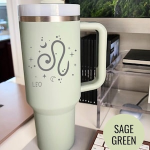 May include: Sage green tumbler with a white lid and handle. The tumbler features a gray Leo zodiac symbol with the word "LEO" printed below. A white straw is inserted into the lid.