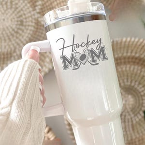 Engraved Hockey Mom tumbler with hockey stick and heart design; stainless steel insulated travel cup with handle; personalized drinkware for sports moms; custom cup for hockey games, practices, or gifts; durable large-capacity tumbler perfect for birthdays, holidays, and hockey season celebrations; stylish engraved design for everyday use.