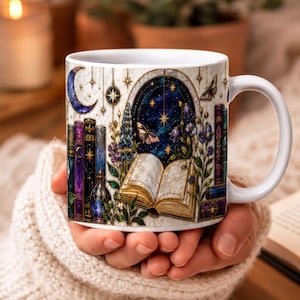 A person's hand holding a white mug with a colorful, whimsical design featuring books, plants, and a starry night sky.